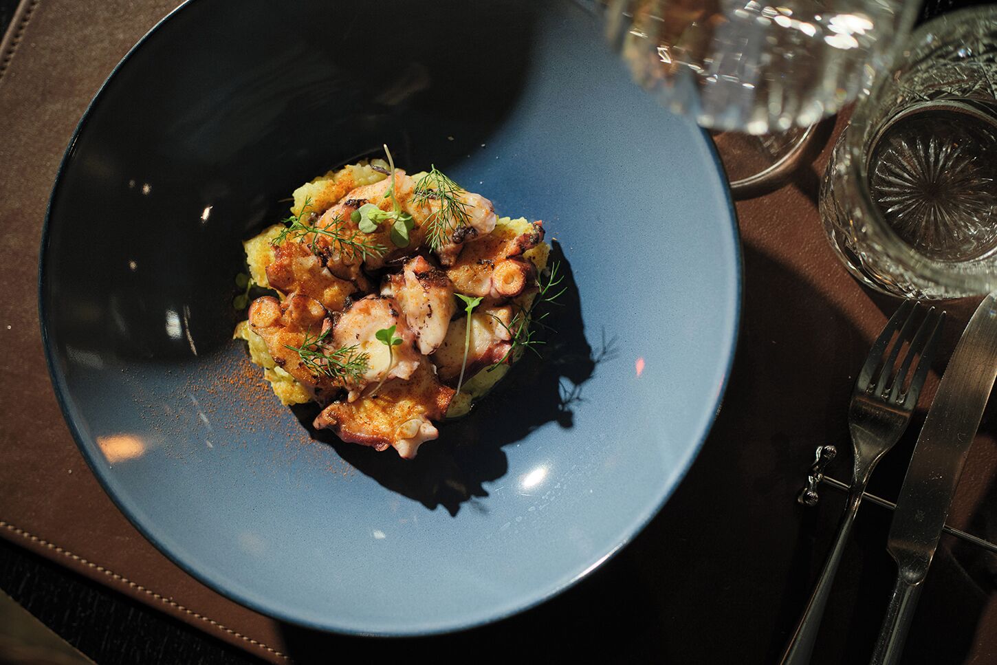 Un plato azul con un plato de mariscos, acompañado de un tenedor y un cuchillo sobre una mesa oscura.