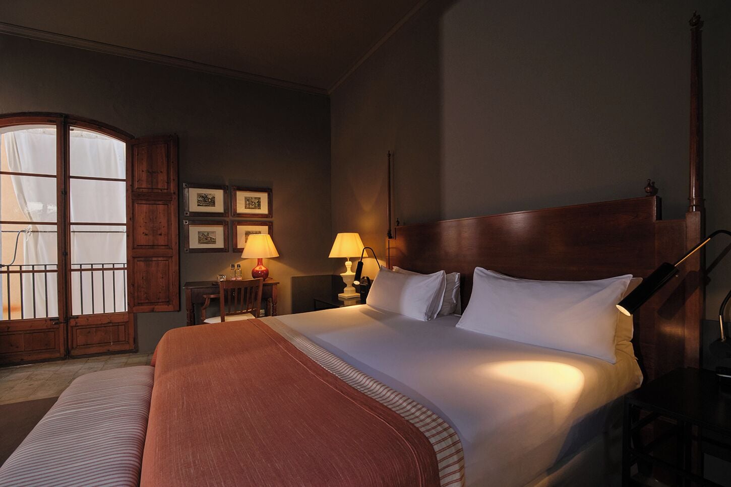 Quarto de hotel com uma cama grande, duas luminárias laterais, um painel de cabeceira em madeira, uma pequena mesa com abajur e uma janela com cortinas.