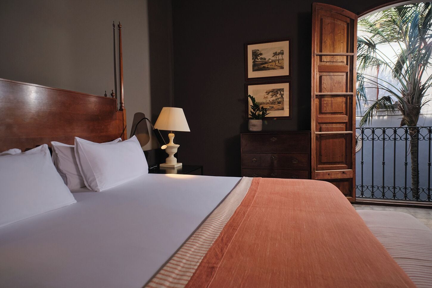 Habitaci ón de hotel con una cama grande, una mesita de noche con lámpara, cómoda y vistas a palmeras a través de un balcón con barandilla de hierro forjado.