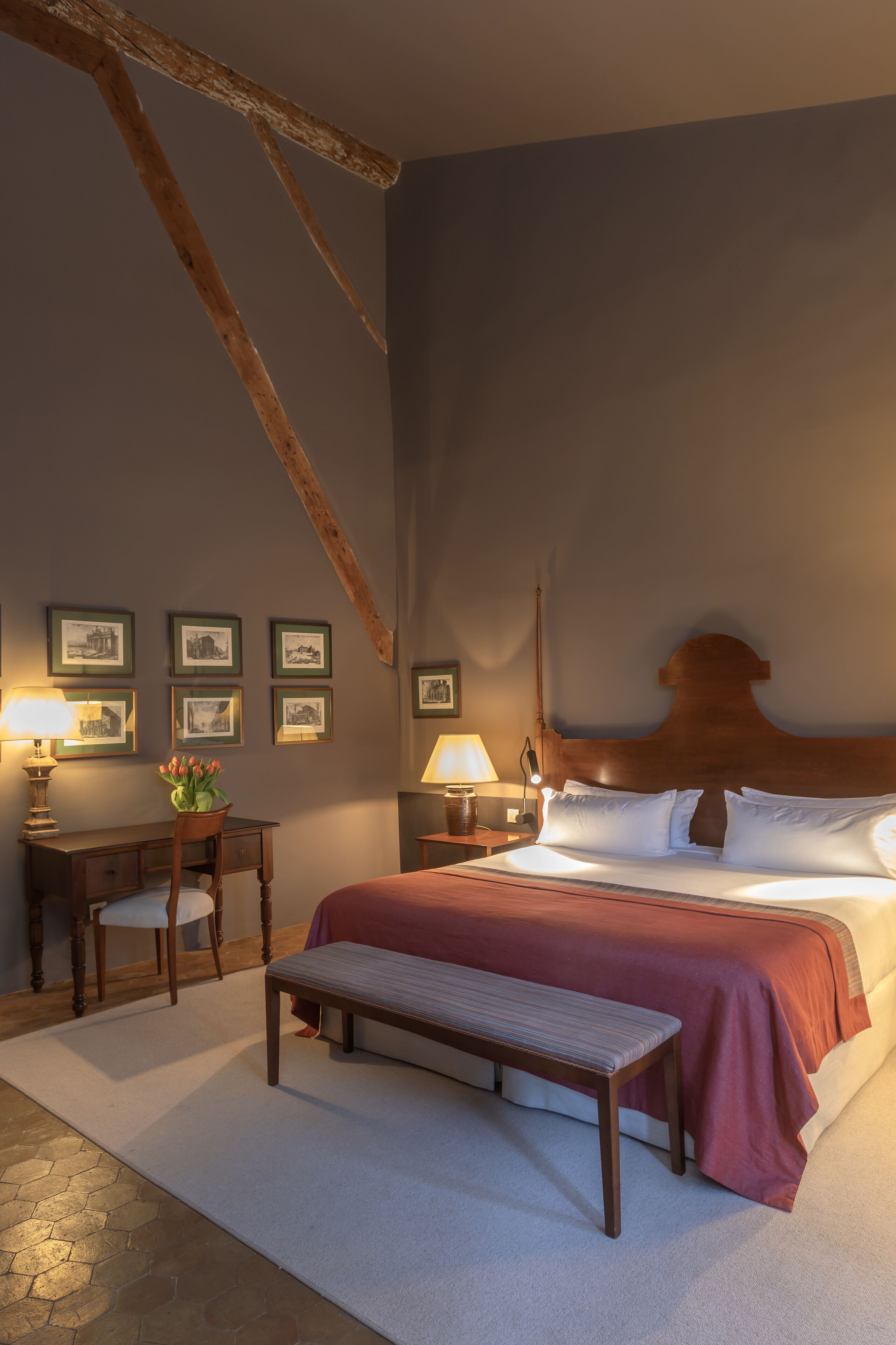 A bedroom with a large bed, a wooden bench, a desk with a chair, and framed pictures on the wall.