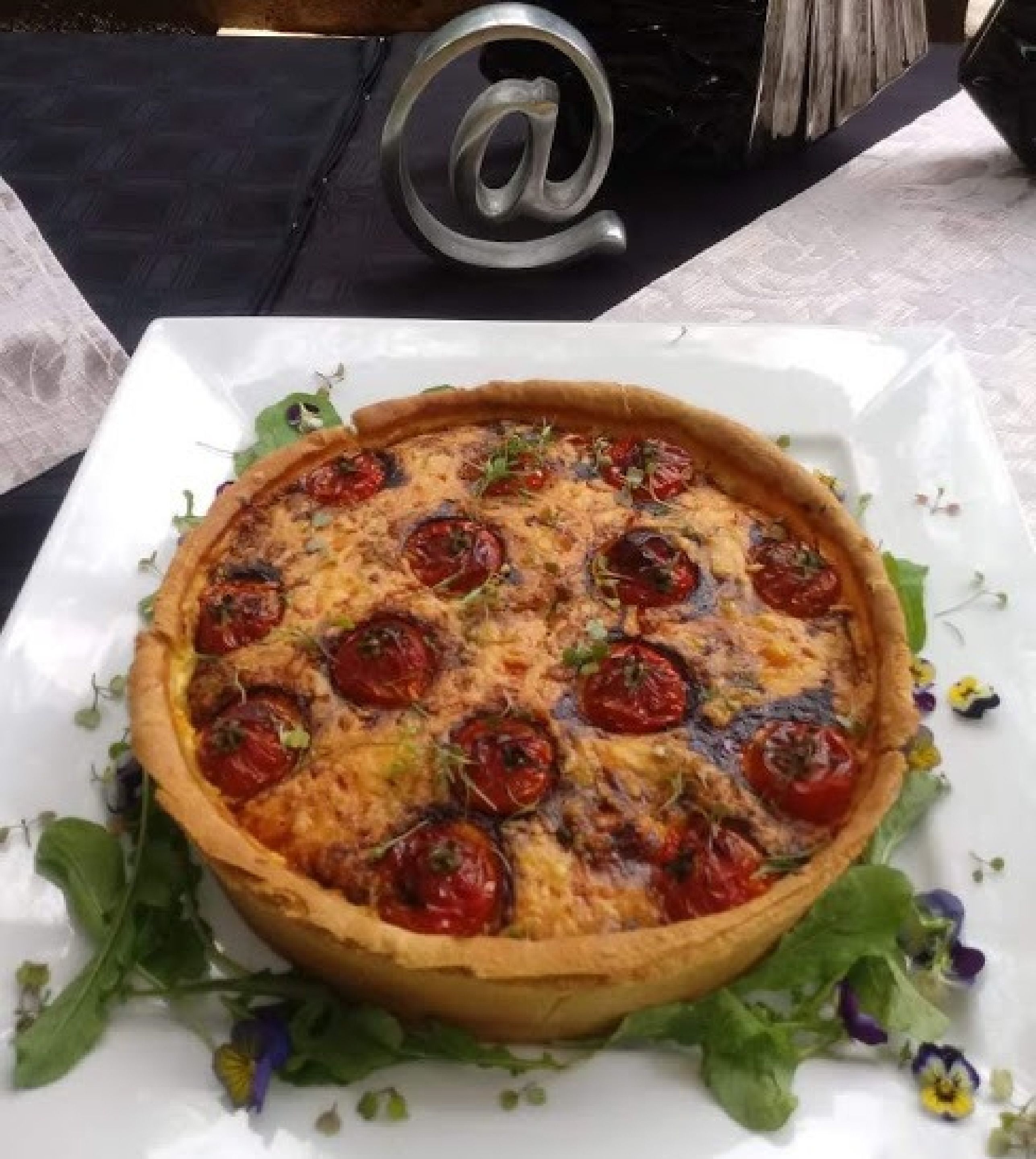 A quiche with cherry tomatoes on a white plate, surrounded by greens and flowers.