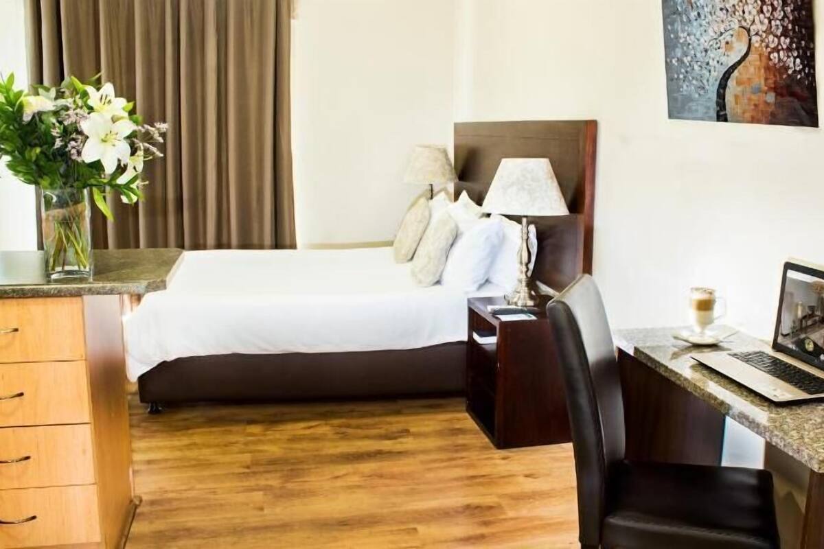 A hotel room with a bed, desk, chair, lamp, laptop, and a painting on the wall.