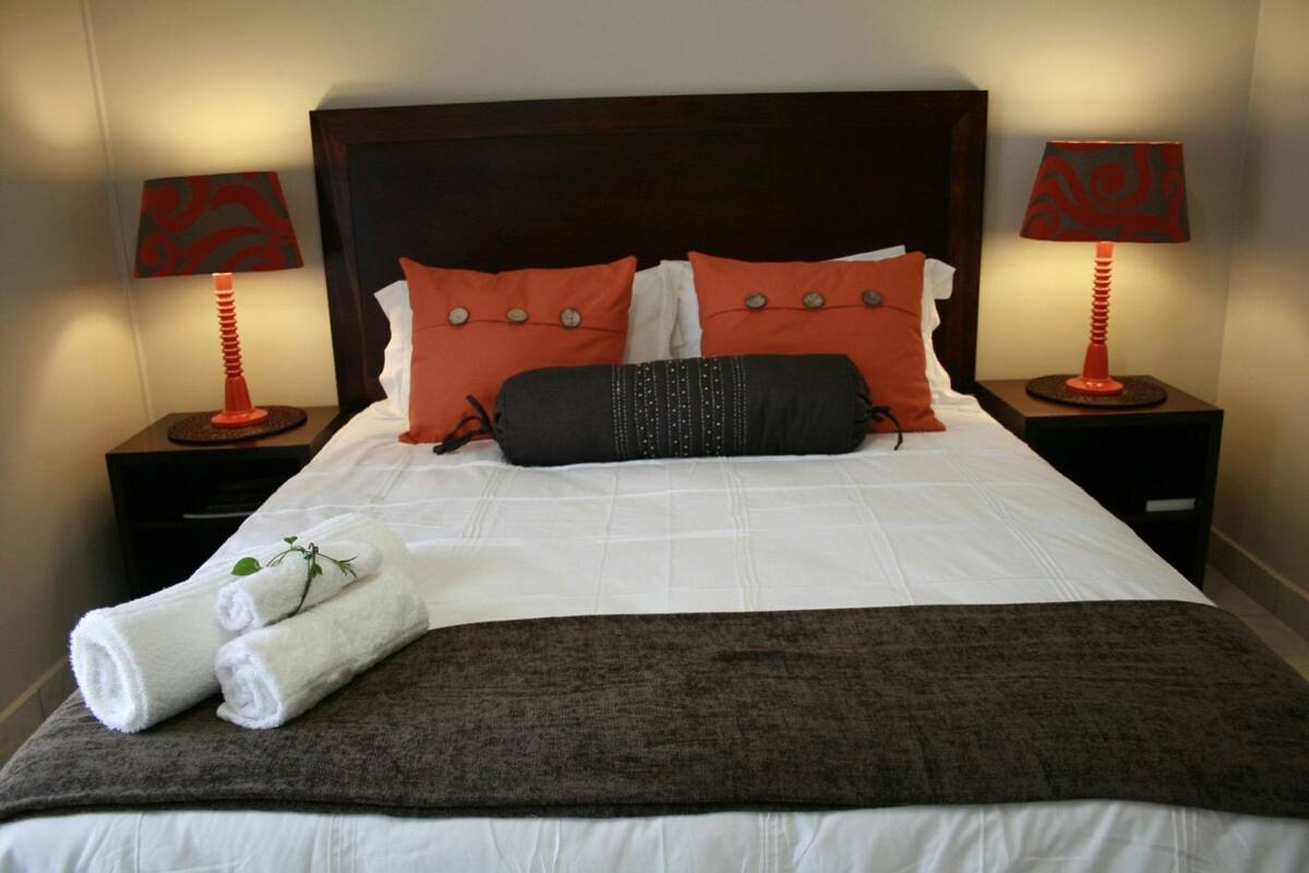 A neatly made bed with white bedding, two orange pillows, a black pillow, and a brown throw blanket. There are two red lamps on nightstands beside the bed.