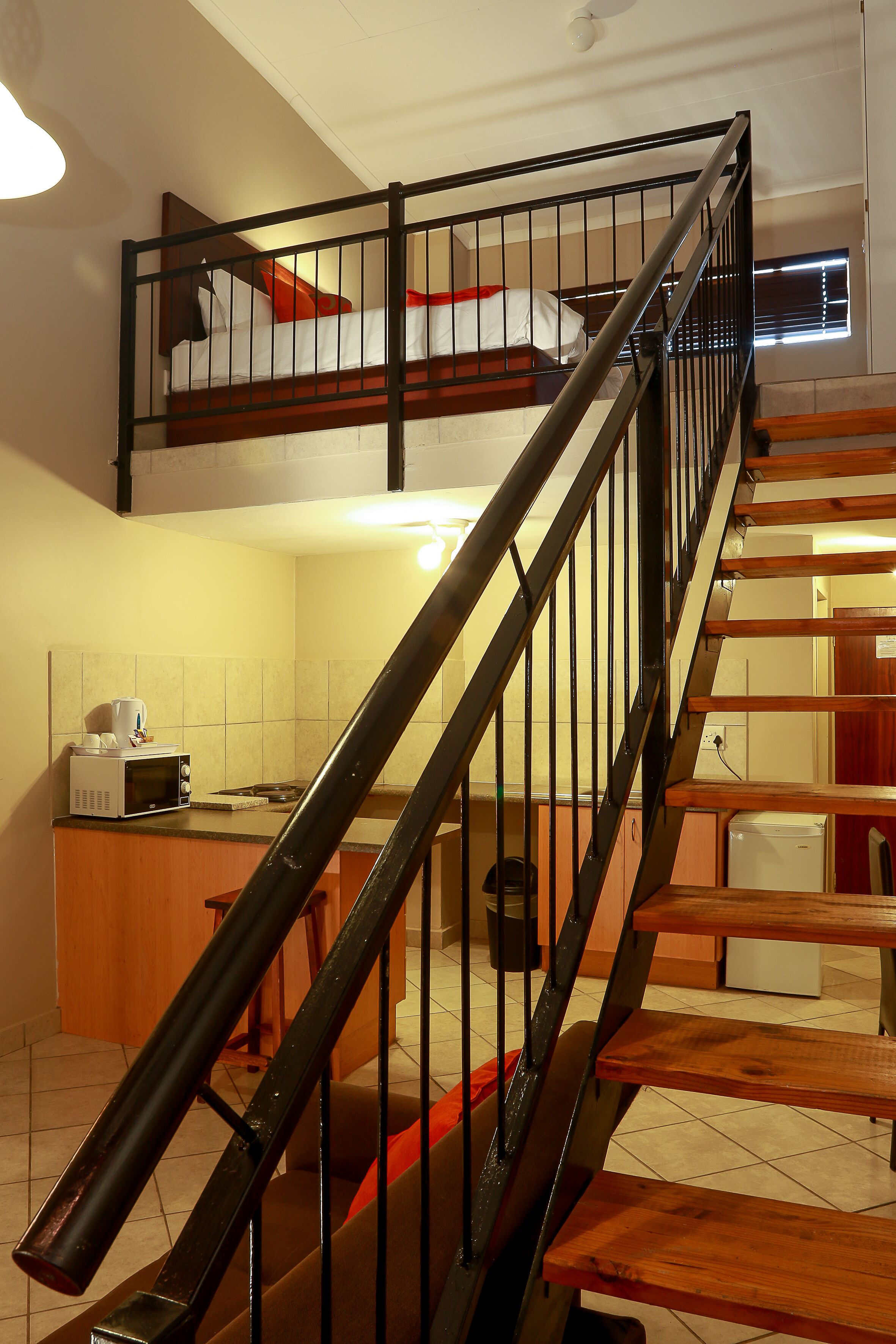 Una escalera con barandilla metálica conduce a un dormitorio en la mezanine con literas. Se ve un microondas y un refrigerador pequeño en la zona de la cocina.