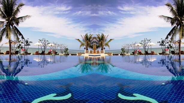 A large, clear blue infinity pool with palm trees and lounge chairs around it.