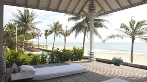 A beachside room with a wooden deck, two beds, and a view of the ocean and palm trees.