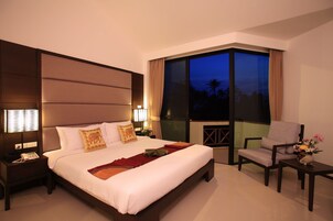 Habitación de hotel con una cama grande, una silla, una mesita con un jarrón y vistas a palmeras a través de un ventanal amplio.