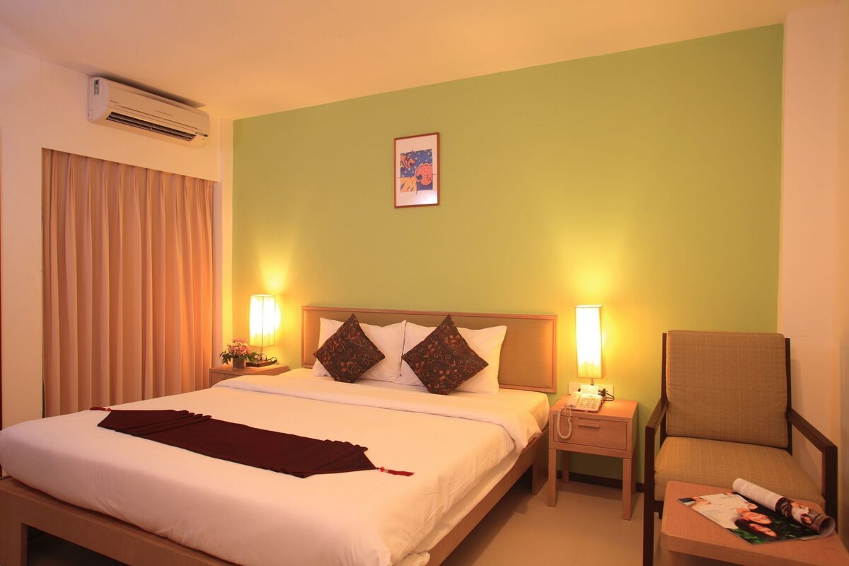 A hotel room with a bed, two bedside lamps, a chair, a nightstand, a phone, and an air conditioning unit.