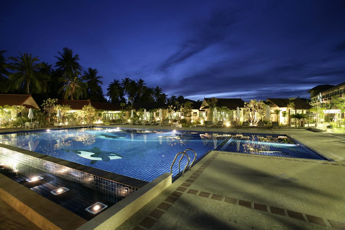 A nighttime swimming pool area with tiled flooring and a clear water surface, surrounded by palm trees and buildings.