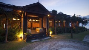 A traditional-style building with a covered outdoor seating area, a dining room, and a well-maintained garden with palm trees.