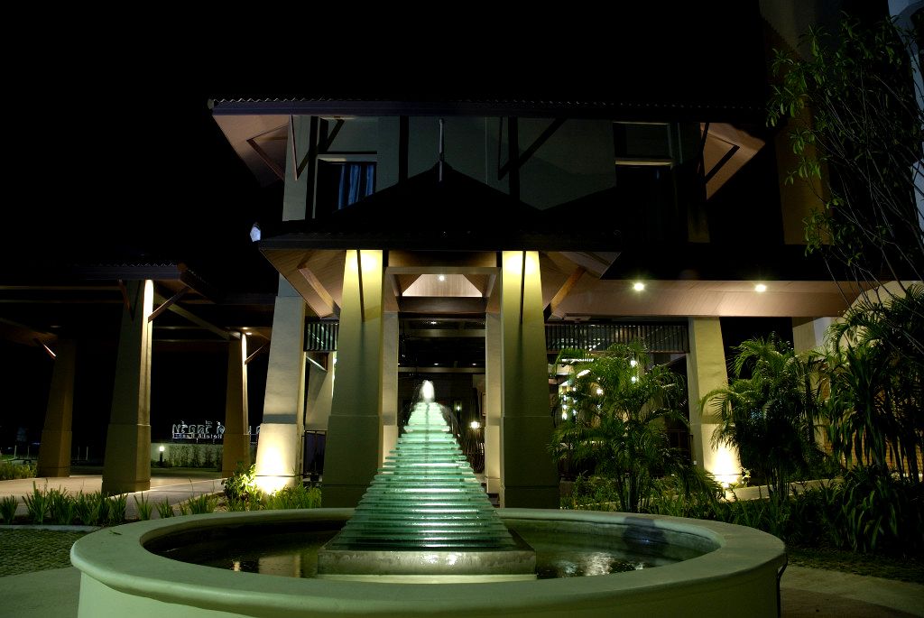 A nighttime view of a building with a central staircase leading to a covered area, surrounded by greenery and a circular water feature.