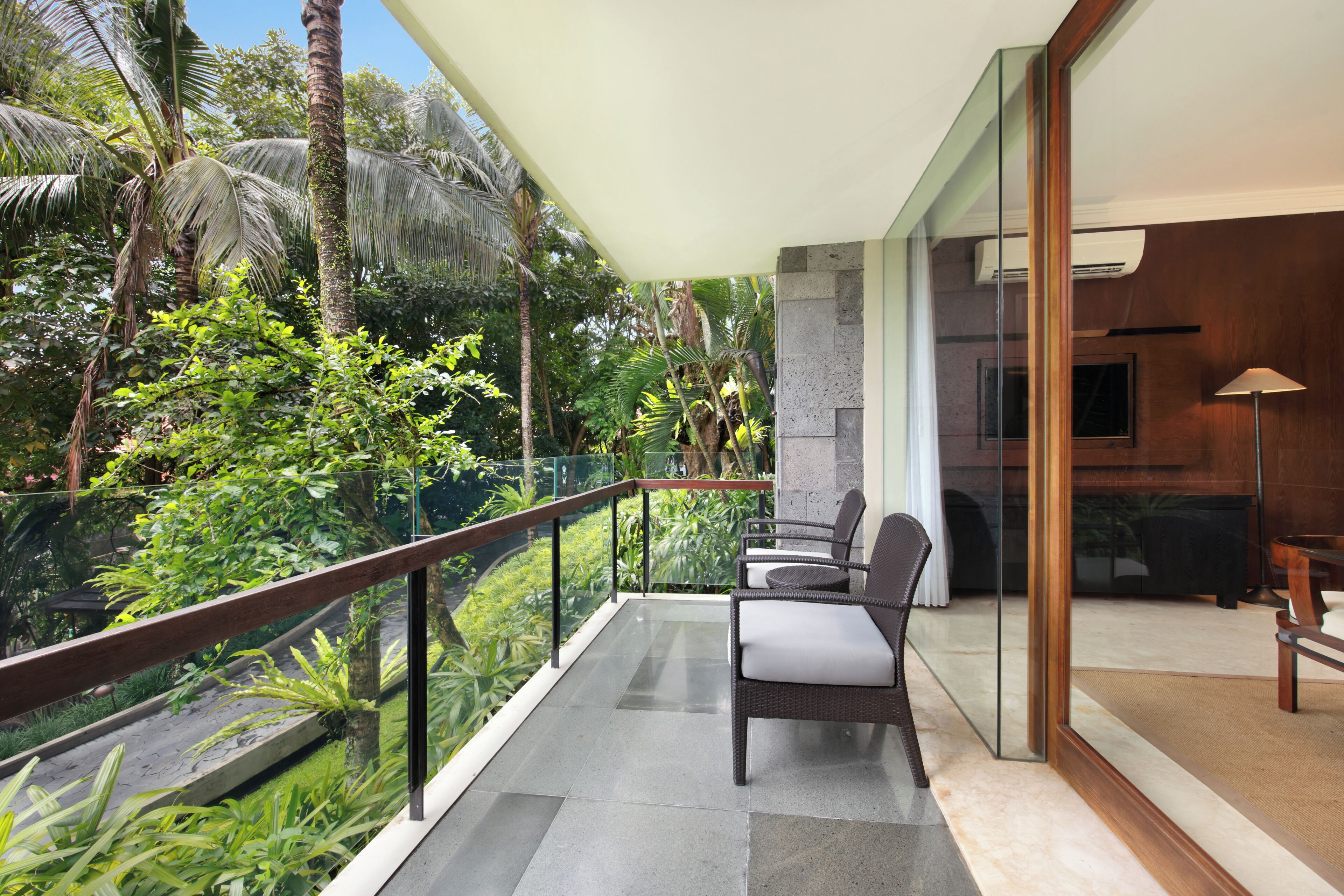 A balcony with a glass railing, a wicker chair, and a view of tropical vegetation.