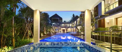 A swimming pool with blue tiles, surrounded by greenery and a building in the background.