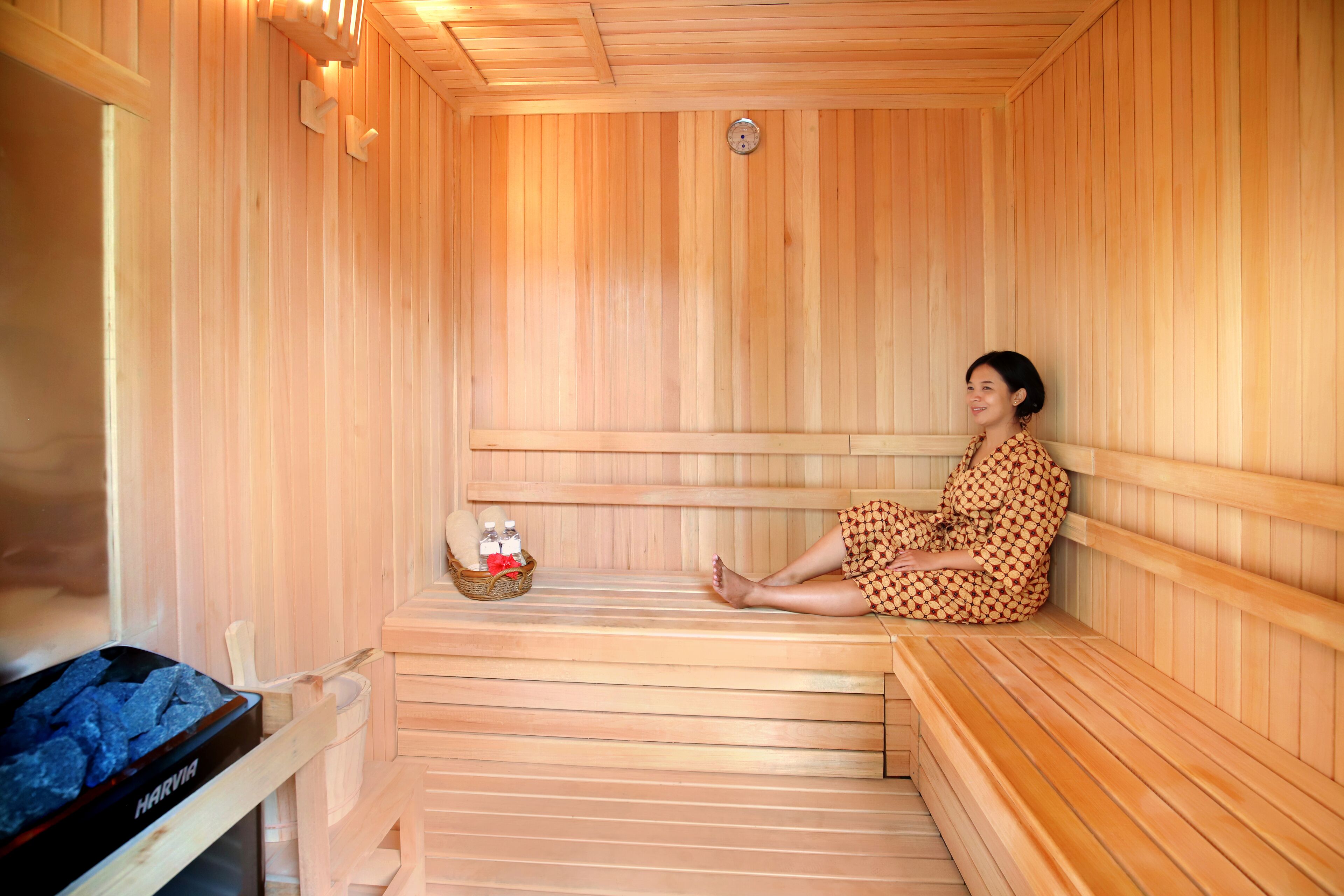 A wooden sauna with a person sitting on a bench, a wooden bench, and a sauna heater.