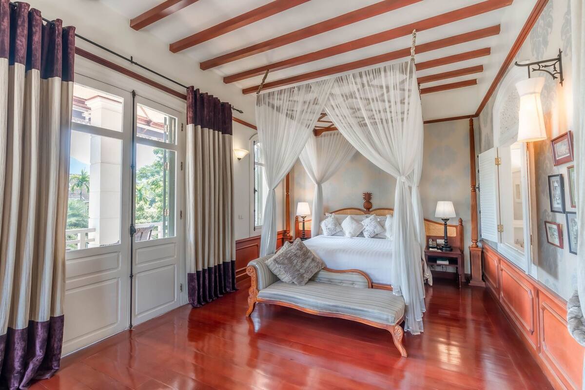 A room with a four-poster bed, a daybed, a wooden floor, and a view through a window.