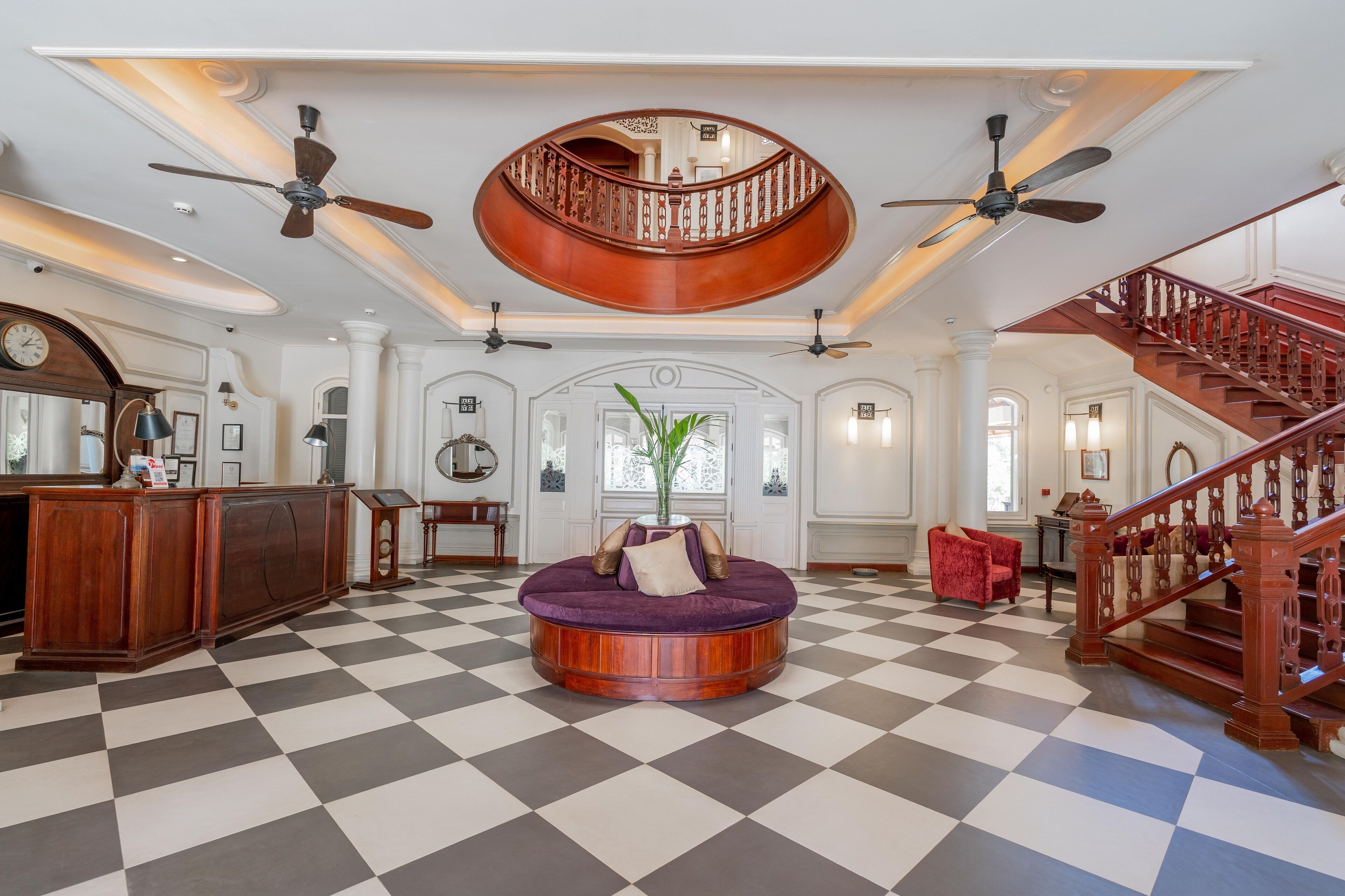 A spacious lobby with a checkered floor, a central round seating area, a staircase with a wooden railing, and a ceiling fan.