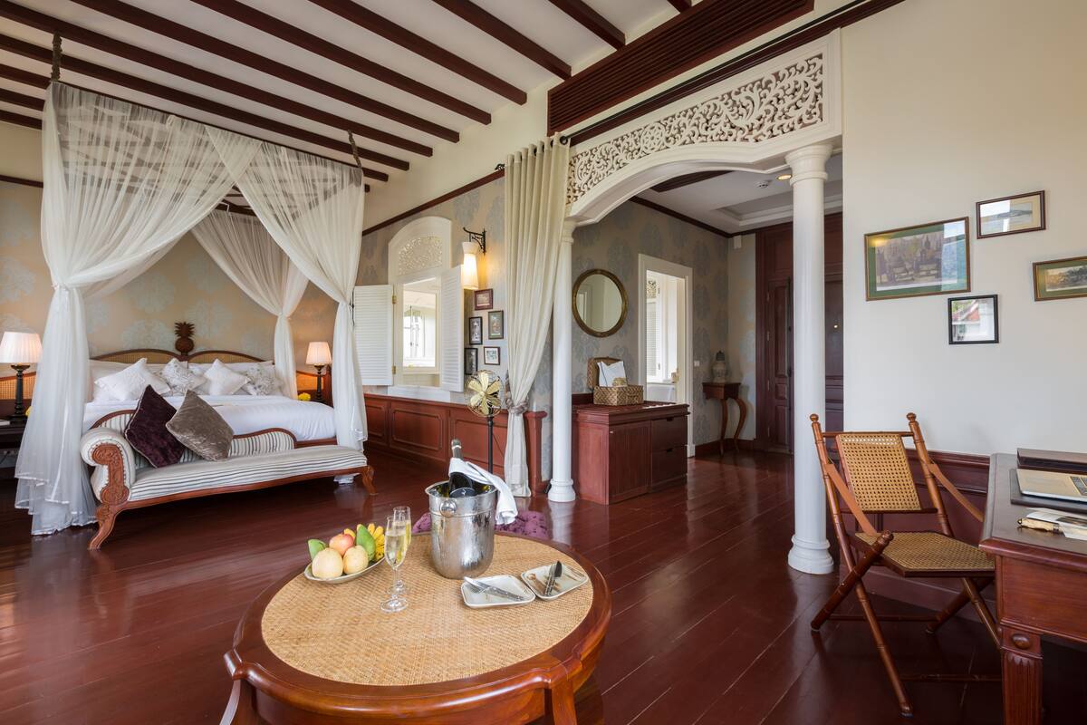A room with a bed, a dining table with fruits and a pitcher, and a chair.