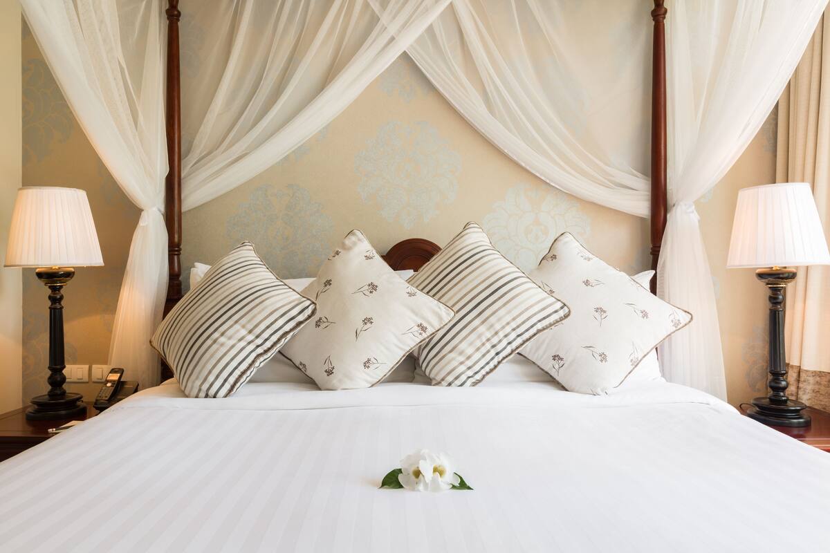 A neatly made bed with white linens and striped pillows, a wooden headboard, and a canopy.