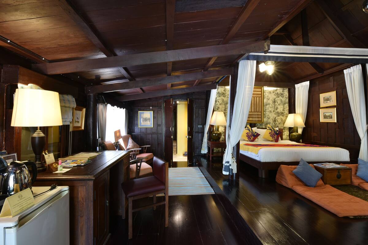 A room with a wooden ceiling, a bed with a canopy, a desk with a lamp, a chair, and a small table with a lamp.