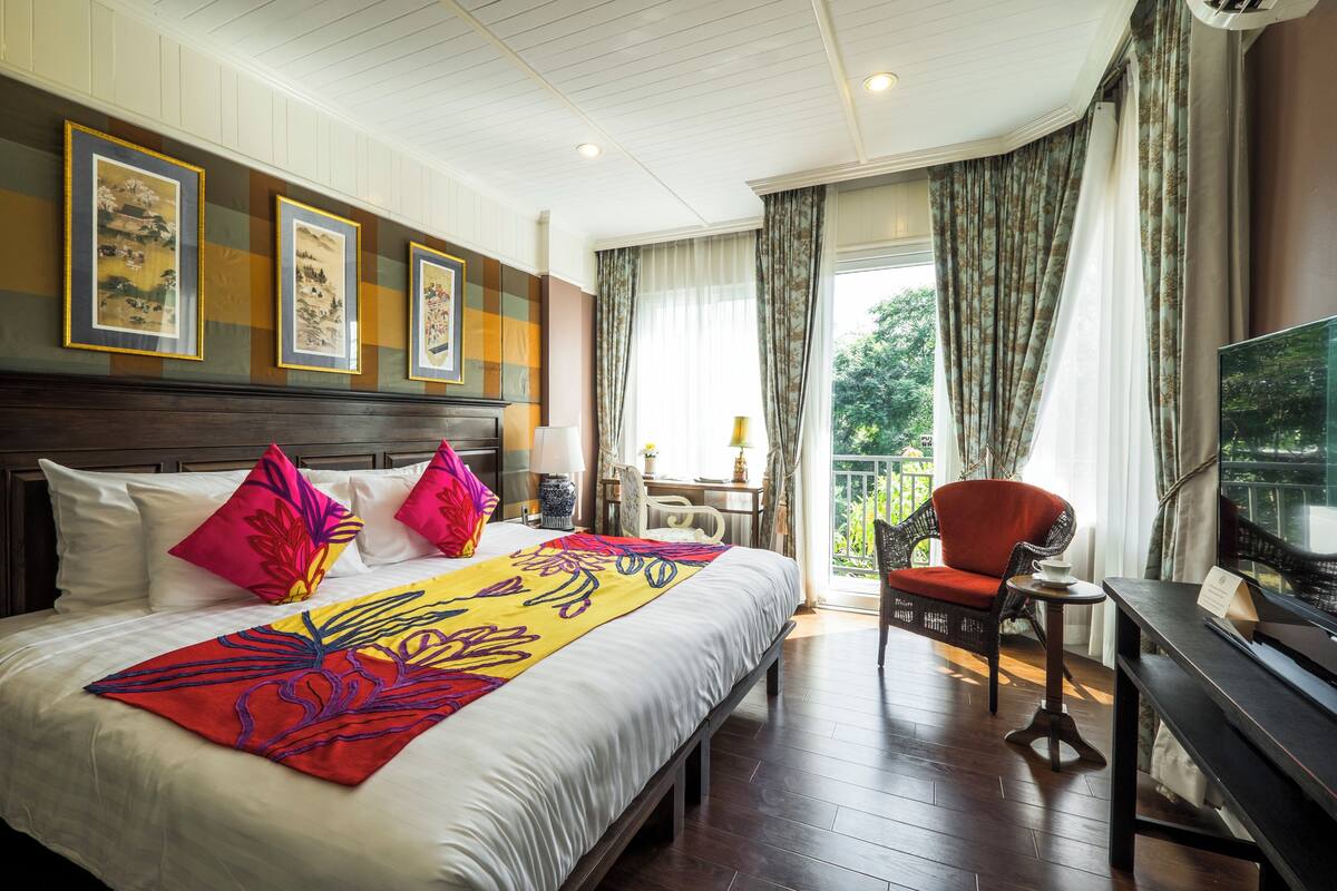 A hotel room with a large bed, a television, a balcony with a chair, and artwork on the wall.