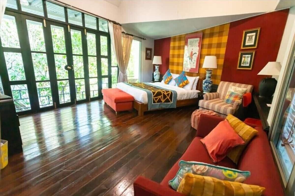 A room with a bed, a sofa, a chair, and a window with a view of greenery.