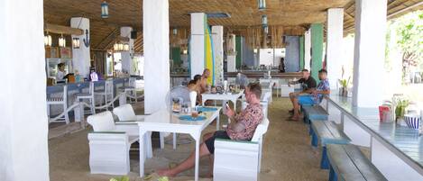 Un restaurante frente al mar con mesas al aire libre, manteles blancos y techo de paja.