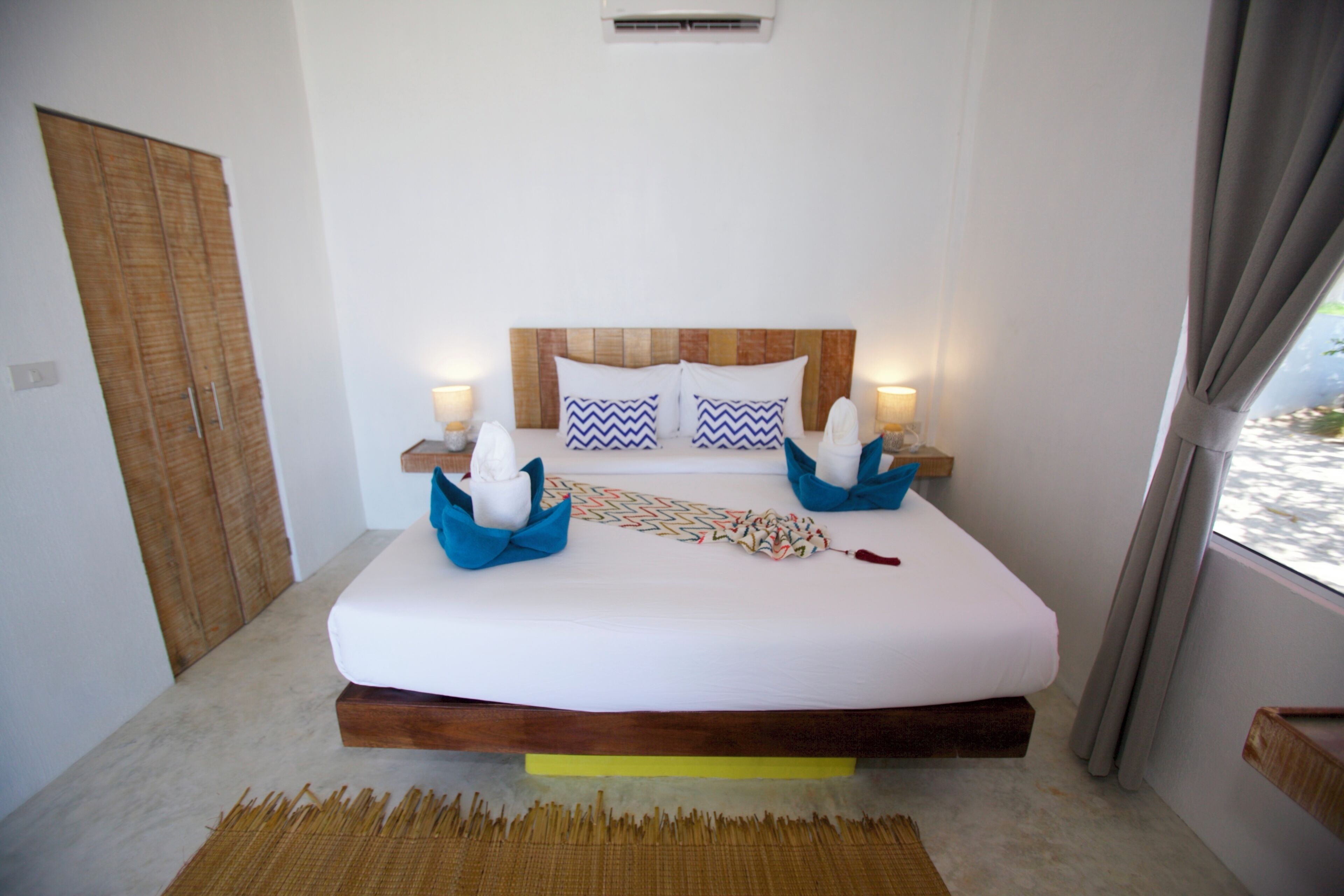Un dormitorio con cabecera de madera, cama con ropa de cama blanca, dos almohadas, lámparas de noche y vistas a través de una ventana.