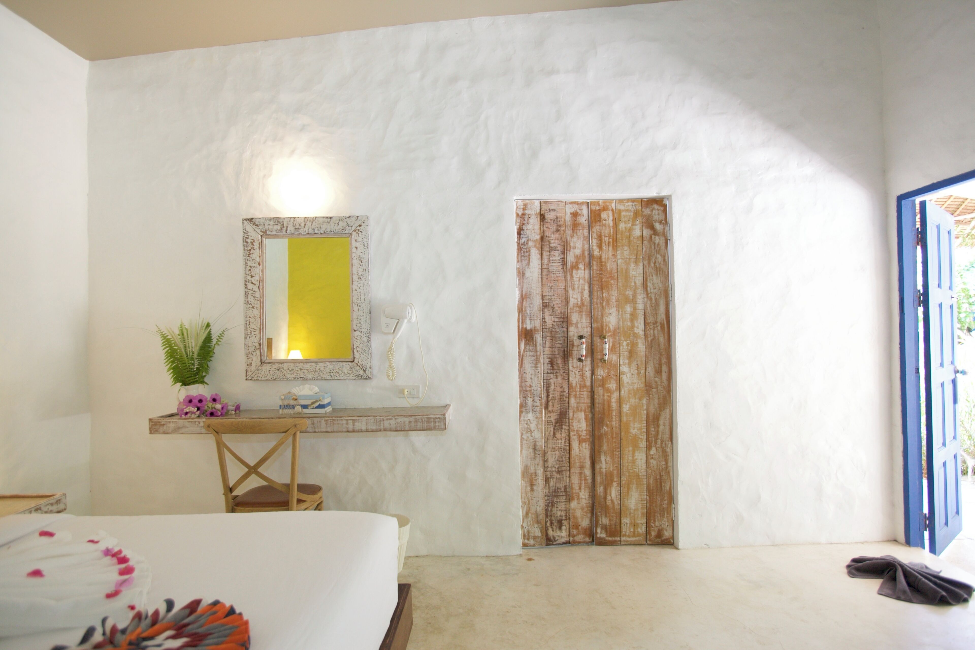 A bedroom with a bed, a desk with a mirror, a plant, and a wooden door.
