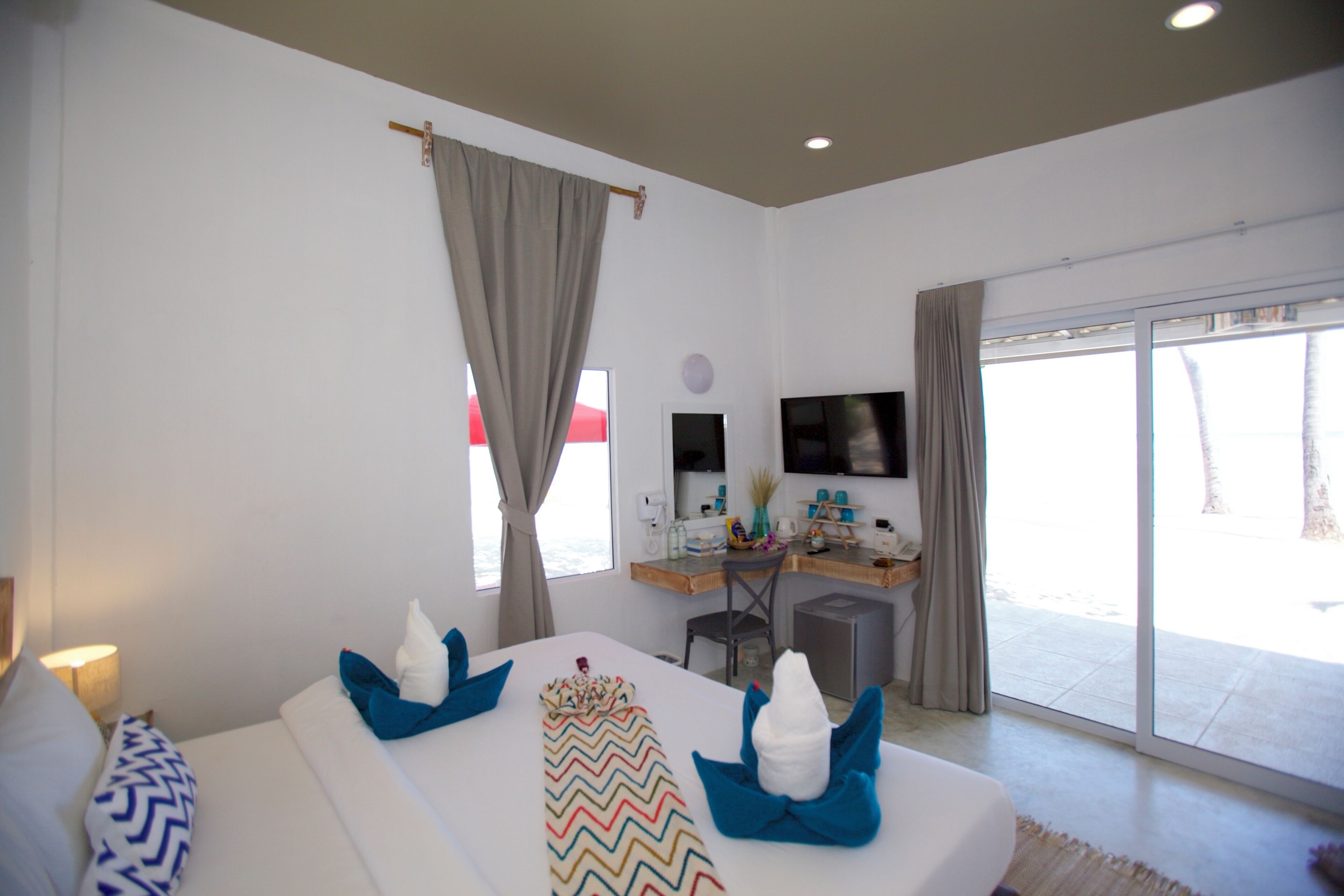 Habitación de hotel con cama, escritorio, silla, televisor y puerta corrediza de vidrio al exterior.