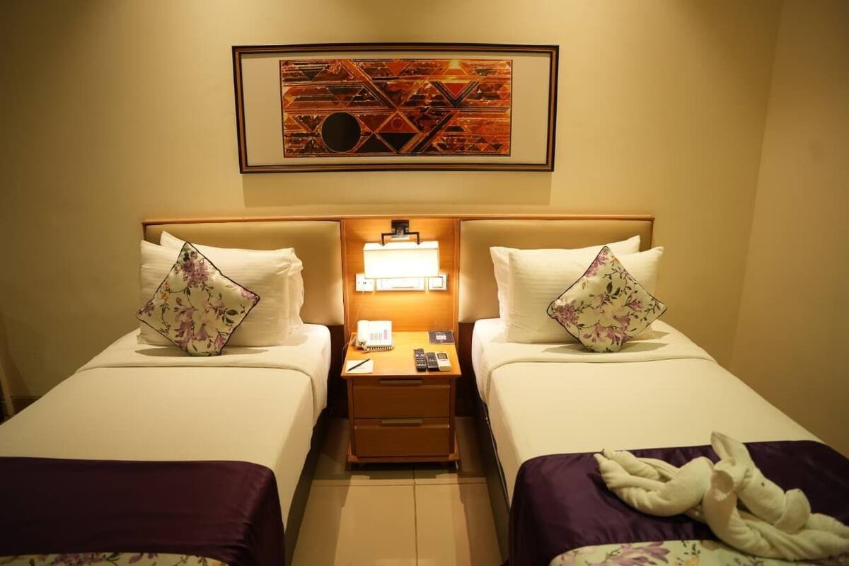 A hotel room with two beds, each with a floral pillow, a bedside table with a lamp, and a framed artwork above the beds.