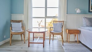 Una habitación con dos sillones de mimbre, una mesita redonda pequeña, una mesita de noche y una cama con cabecera blanca.