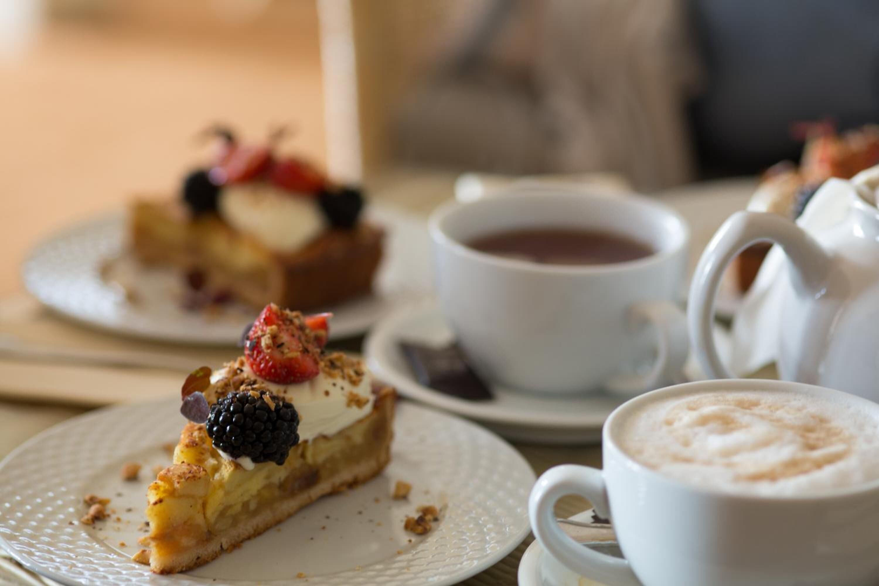 Un plato de postre con una porción de tarta cubierta de bayas y crema batida, una taza de café y una tetera sobre una mesa.