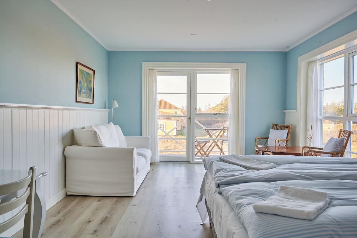 A bedroom with a white bed, a white armchair, a dining table with chairs, and a large window with a view of the outside.