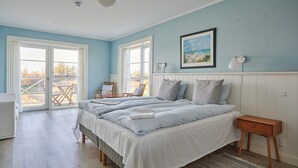 Un dormitorio con una cama grande, dos ventanas, un cuadro, una mesita de noche y una silla junto a la ventana.