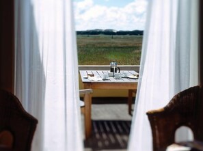 A view through a window with curtains, revealing a wooden table with cups and a coffee pot, set on a deck with a grassy field and hills in the background. - Hjorths Badehotel (Skagen)