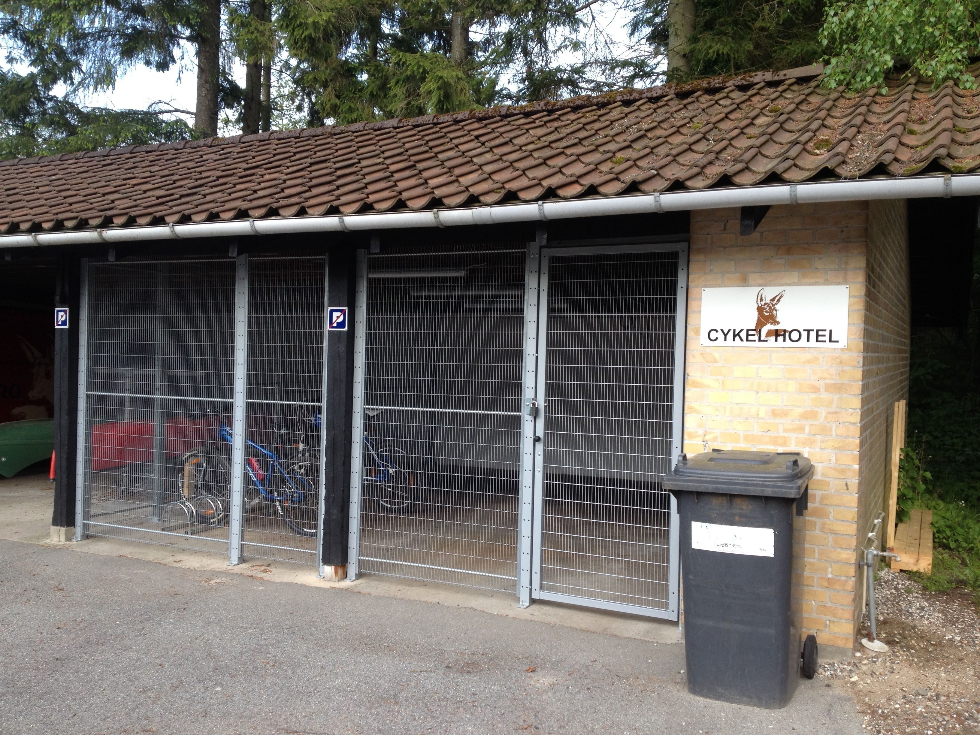 A building with a tiled roof, a sign reading "CYKEL HOTEL", and a fenced parking area with bicycles.