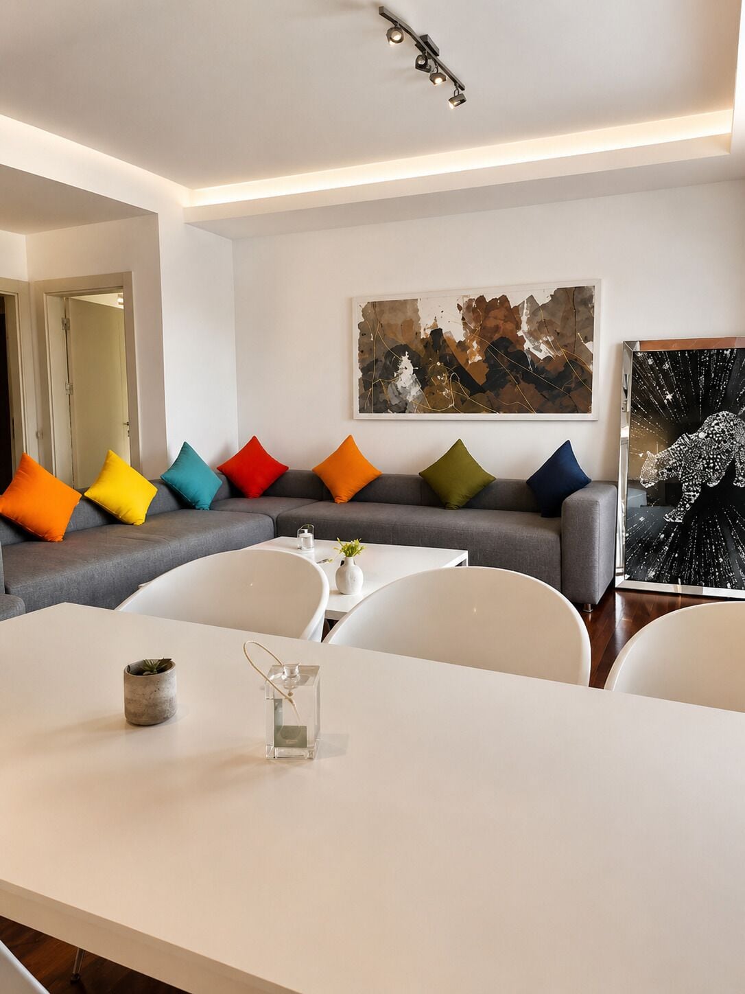 Ein modernes Wohnzimmer mit einem grauen Ecksofa, bunten Kissen, einem weißen Esstisch und einem großen, abstrakten Gemälde an der Wand.