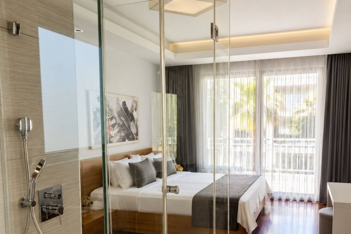 Ein modernes Hotelzimmer mit einer in Glas eingefassten Dusche, einem Bett in Grau-Wei ß gehalten, einem Nachttisch und einem großen Fenster mit Vorhängen.