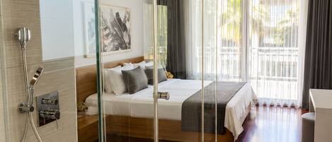 Una habitación de hotel moderna con ducha de vidrio, cama con decoración en gris y blanco, una mesita de noche y un amplio ventanal con cortinas.