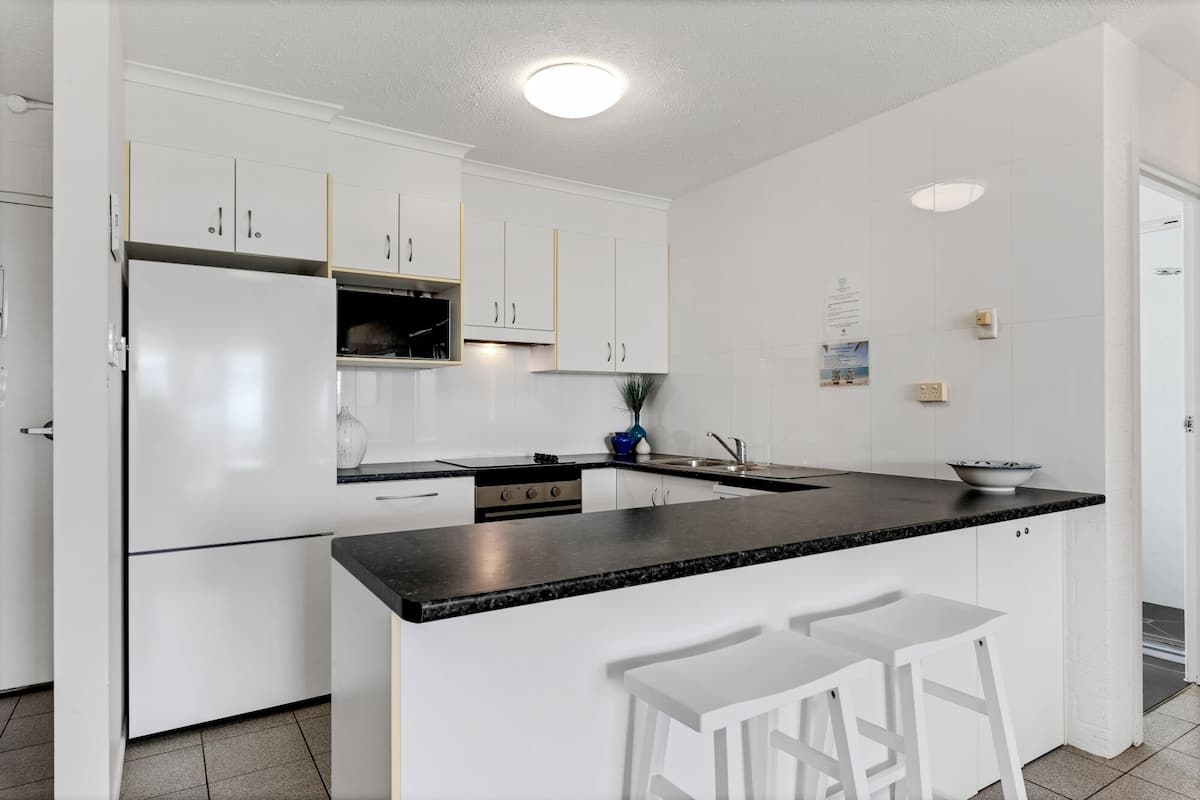 A modern kitchen with white cabinets, a black countertop, and a built-in oven. There are two white bar stools at the island.