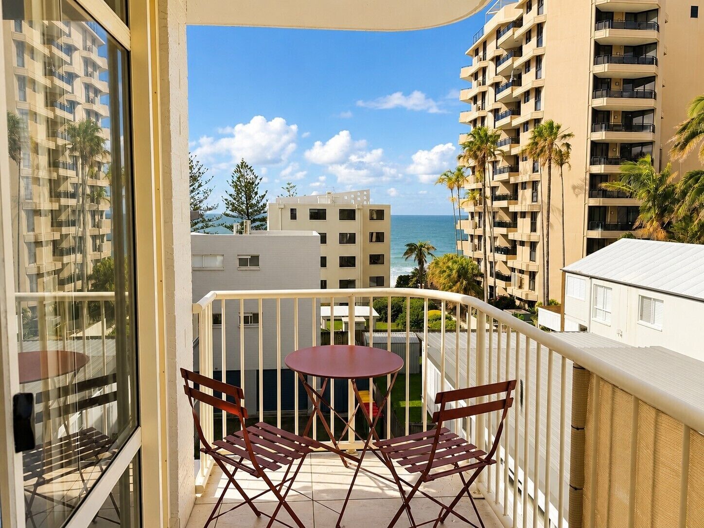 Un balcon avec une table et des chaises, donnant sur une zone côtière avec des bâtiments et des palmiers.