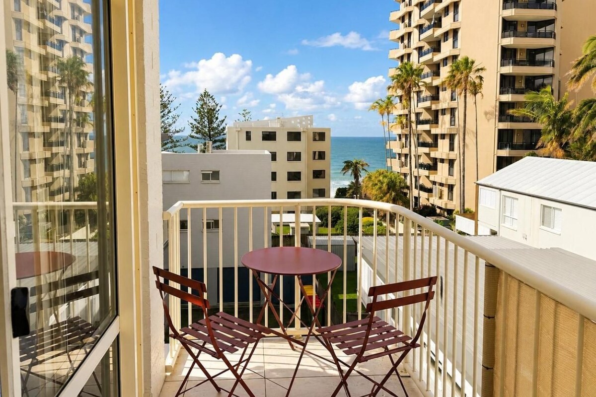 Un balcon avec une table et des chaises, donnant sur un quartier résidentiel et l’océan.
