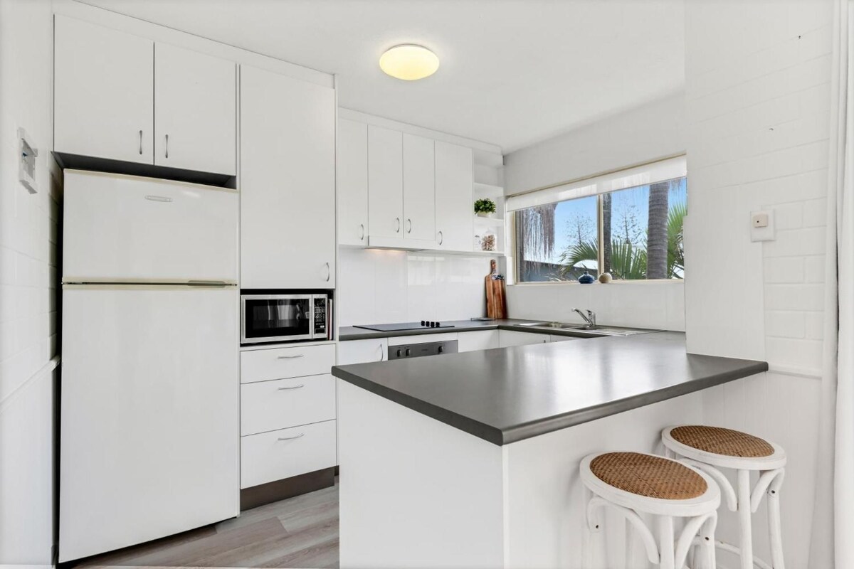 A modern kitchen with white cabinetry, a stainless steel island, and two wicker stools.