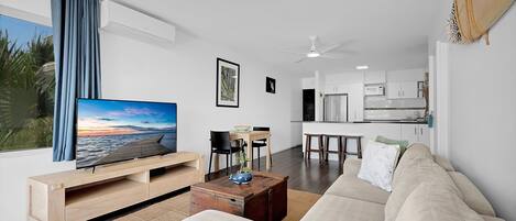 A modern living room with a large flat-screen TV, a sofa, a coffee table, and a dining area with chairs and a small table.