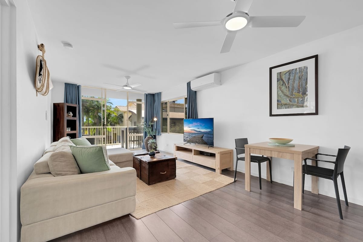 A modern living room with a sofa, coffee table, TV, dining table, chairs, and a ceiling fan.