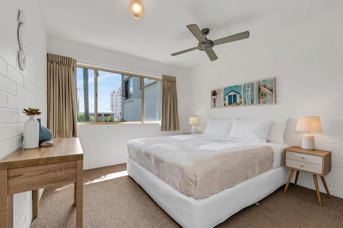 A hotel room with a bed, bedside tables, a desk, a ceiling fan, a window with curtains, and a wall-mounted artwork.