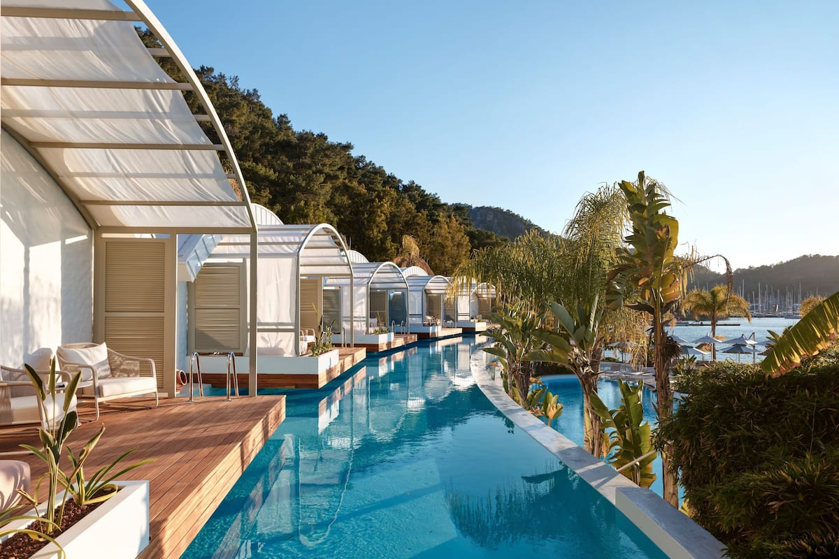 A poolside area with sun loungers and cabanas, surrounded by tropical plants and overlooking a body of water with boats in the distance.