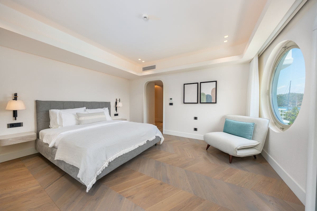 A modern bedroom with a large bed, a bedside table with a lamp, a window with a view, and a small white chair.