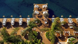 An aerial view of a wooden deck with multiple tables and chairs, surrounded by lush greenery and overlooking a body of water.