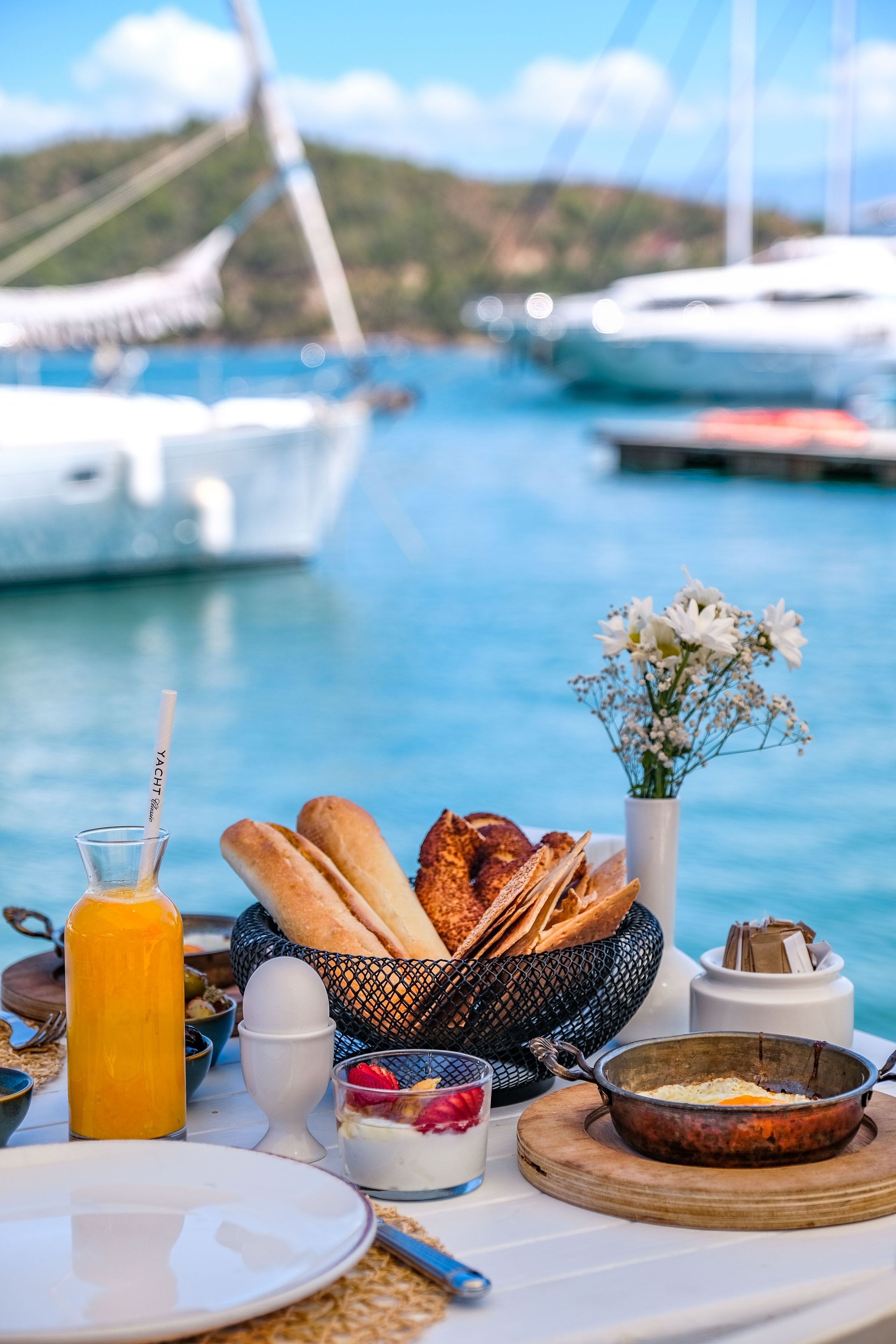 A breakfast table with bread, eggs, and a fruit bowl by a marina with boats in the background.